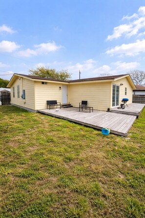 A single-story yellow house with a covered patio area, a barbecue grill, and two chairs.