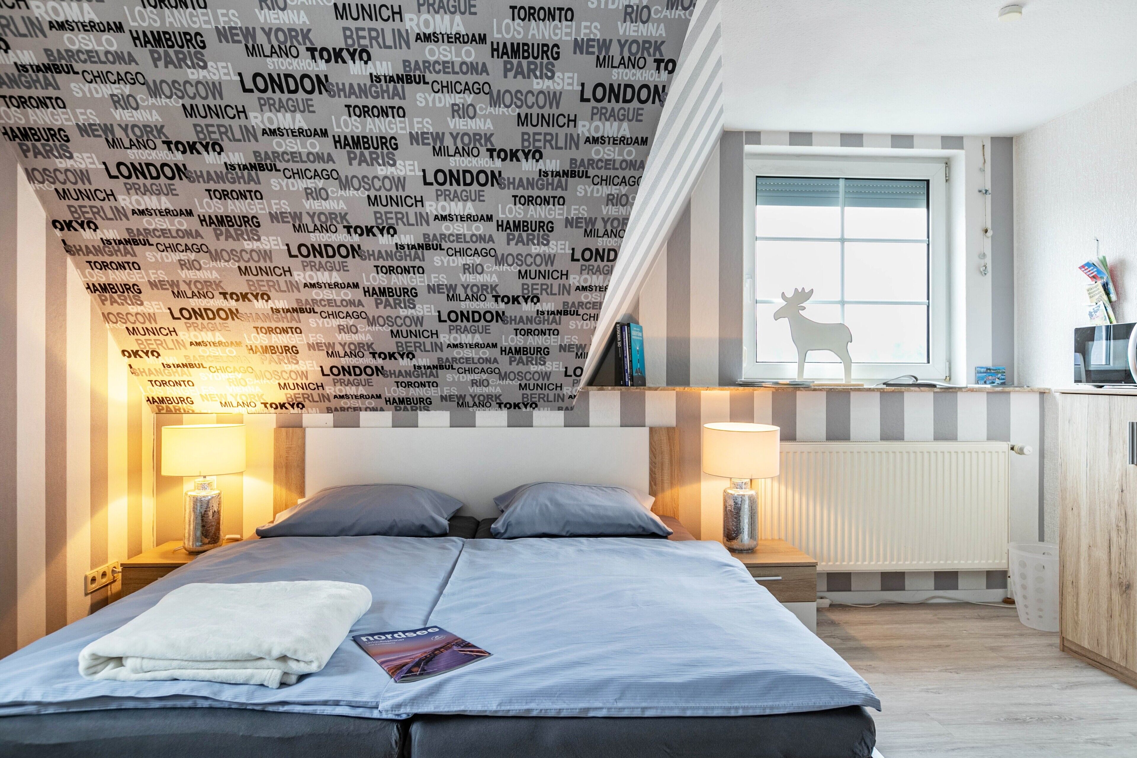 A hotel room with a bed, two bedside lamps, a window with a view, and a wall with city names.