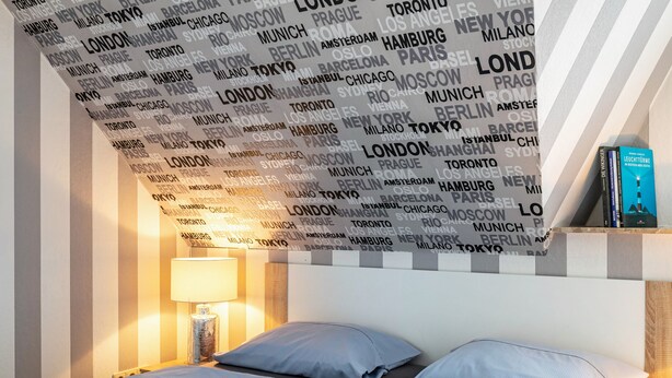 Habitación de hotel con cama, almohadas, mesita de noche con lámpara y una pared con nombres de ciudades.