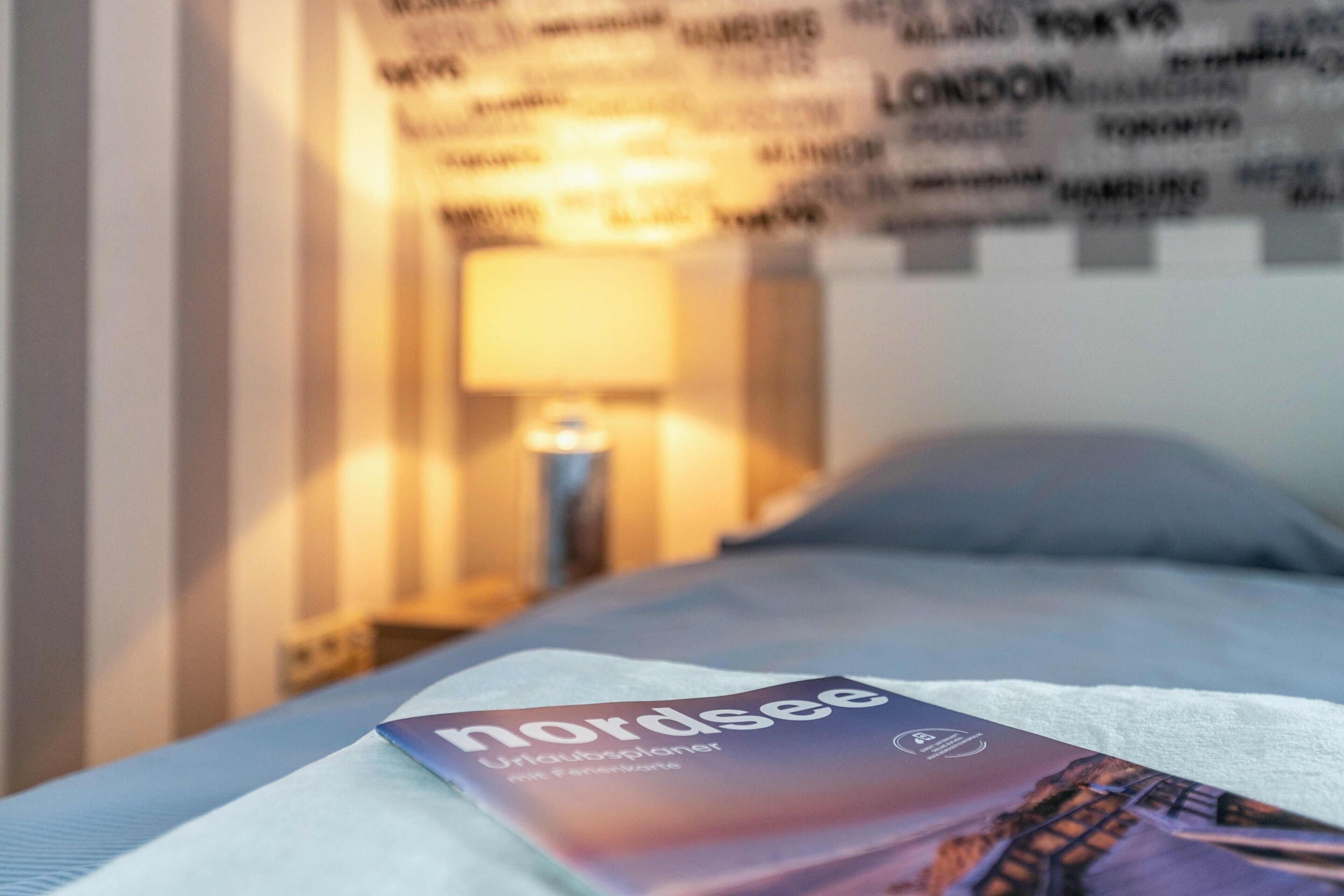 Un letto con una brochure Nordsee, una lampada e un muro con nomi di città.