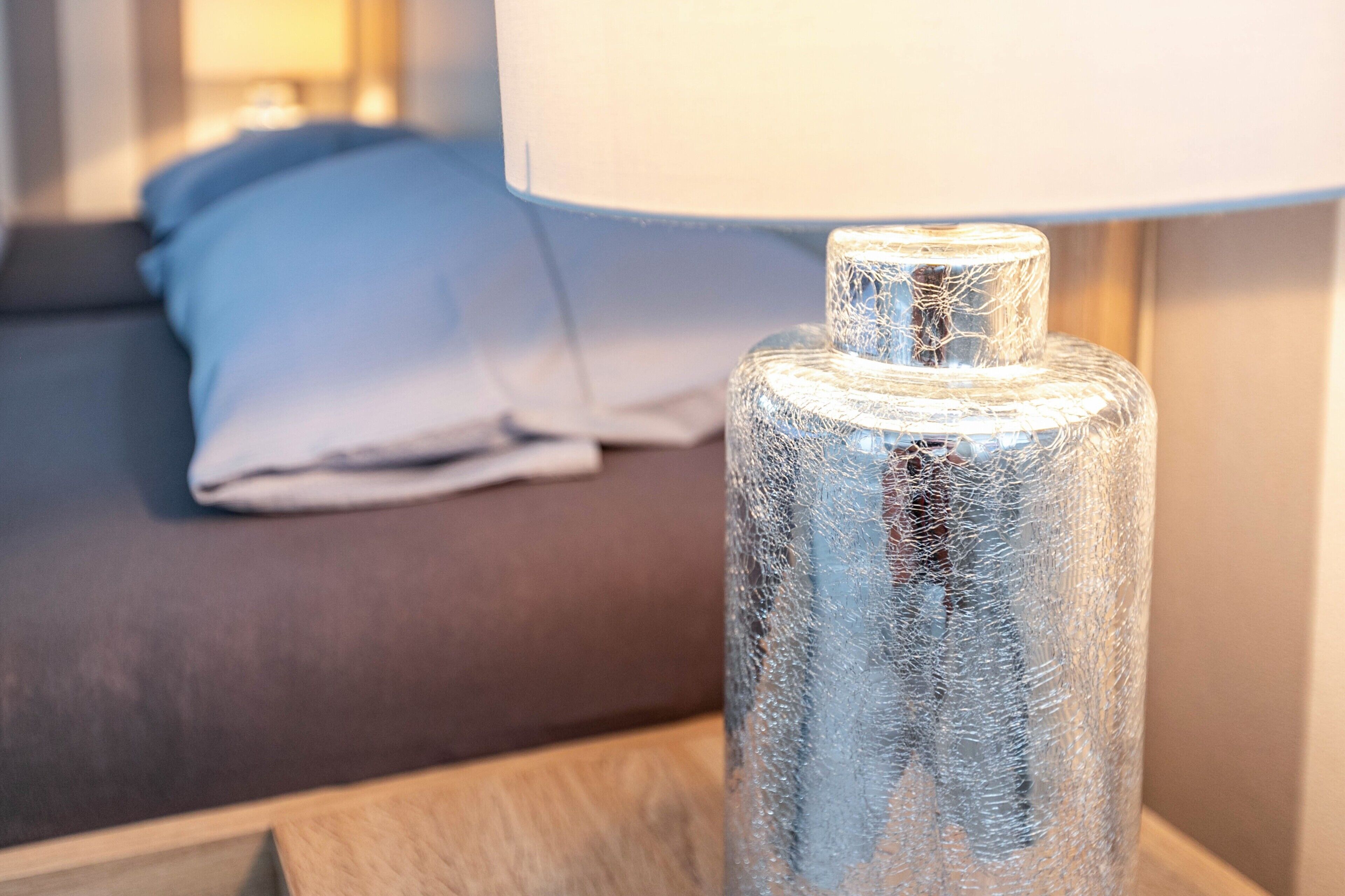 En tydlig bild av en lampa med strukturerat glas, placerad på ett nattduksbord. I bakgrunden syns en säng och en annan lampa.