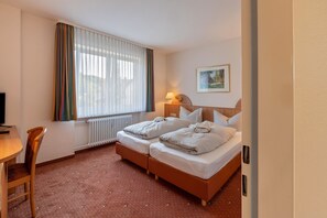 Habitación de hotel con dos camas, un escritorio, una silla, una lámpara y un cuadro en la pared.