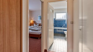 Une chambre d’hôtel avec un lit, une table de chevet avec une lampe, et une vue sur une salle de bain dont le mur est recouvert de carreaux bleus et qui contient des toilettes blanches.