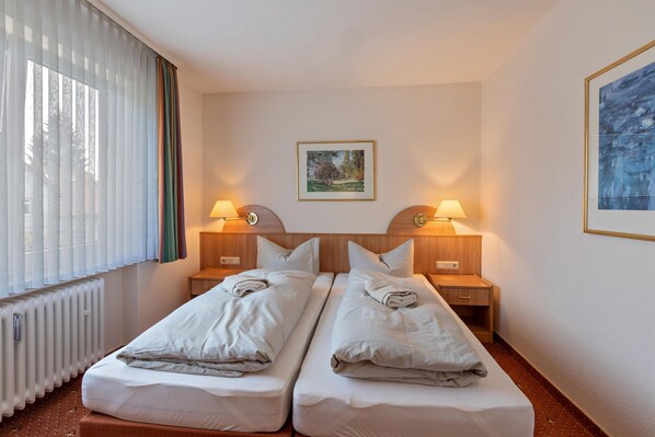 Una habitación de hotel con dos camas individuales, un radiador, dos mesitas de noche con lámparas, un cuadro enmarcado en la pared y una ventana con persianas.