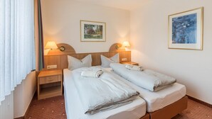 Ein Hotelzimmer mit zwei Betten, einem Nachttisch mit Lampe, einer kleinen Schublade und zwei gerahmten Bildern an der Wand.