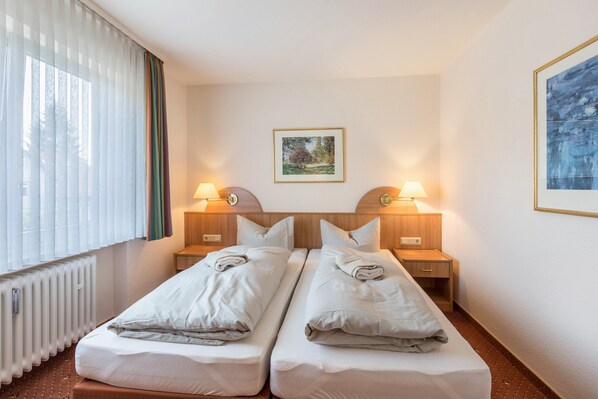 Habitación de hotel con dos camas individuales, un radiador, una ventana con persianas y dos lámparas de noche, además de un cuadro en la pared.