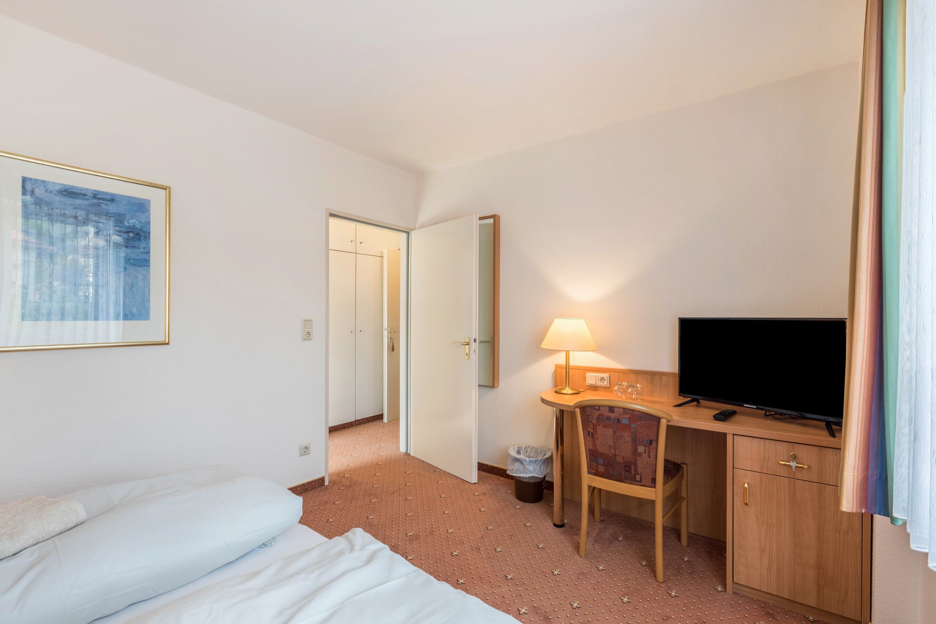 Habitación de hotel con una cama, un escritorio con televisor, una silla, un cubo de basura y una puerta que da a otra habitación.