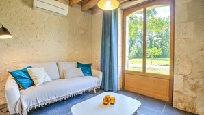 A room with a white sofa, a small table with three glasses, and a view of a garden through a large window. - Gîte Le Pigeonnier with Shared Pool, Wi-Fi, and Air Conditioning (Chassaignes)