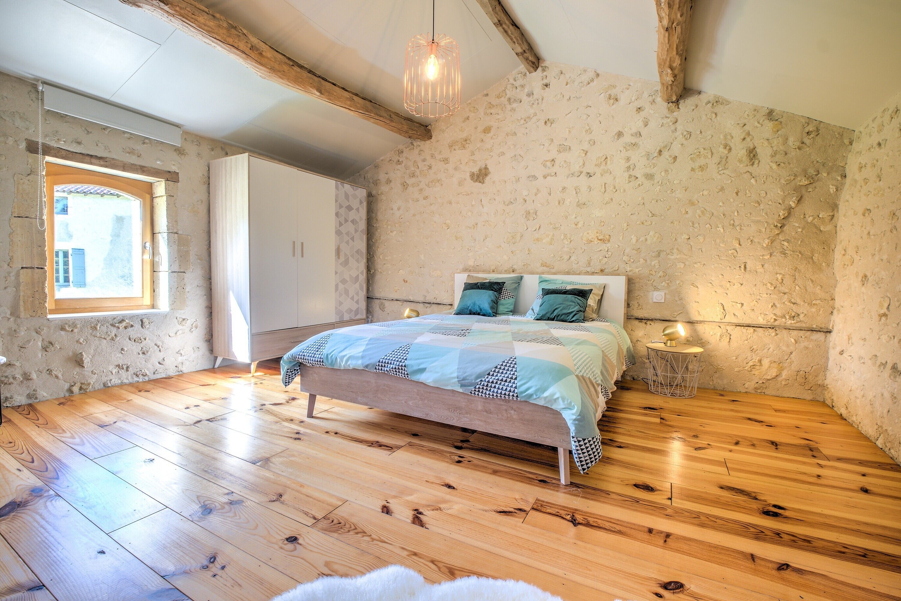 Een slaapkamer met een houten vloer, een bed met een beddengoed met een patroon, een hanglamp en een stenen muur.