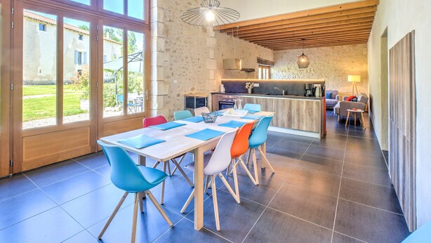 Un vaste espace dédié à la salle à manger, avec une grande table et des chaises aux couleurs vives, une cuisine en arrière-plan et une vue sur l’extérieur.