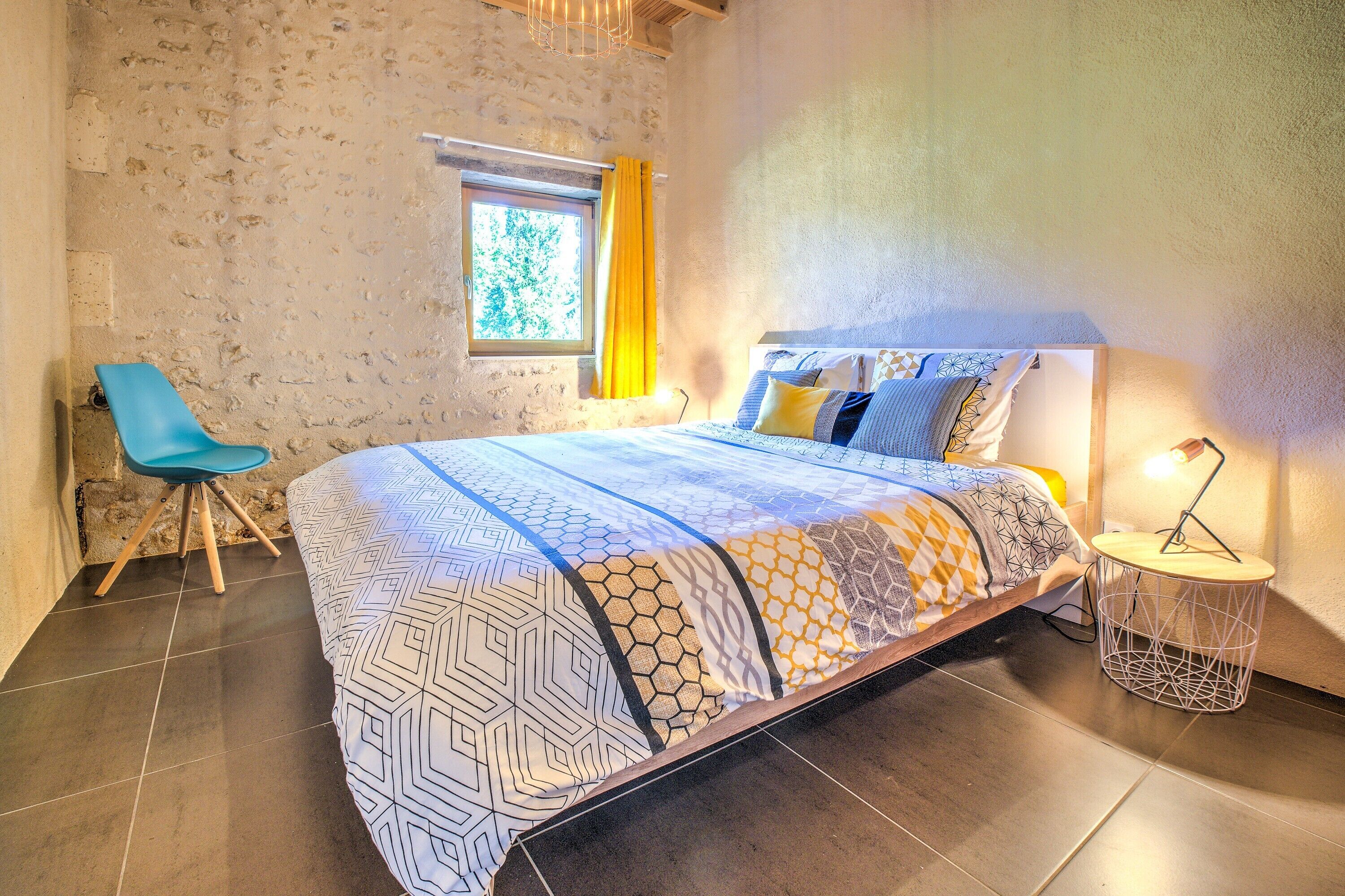 A bedroom with a bed, a chair, a window with curtains, and a bedside lamp.