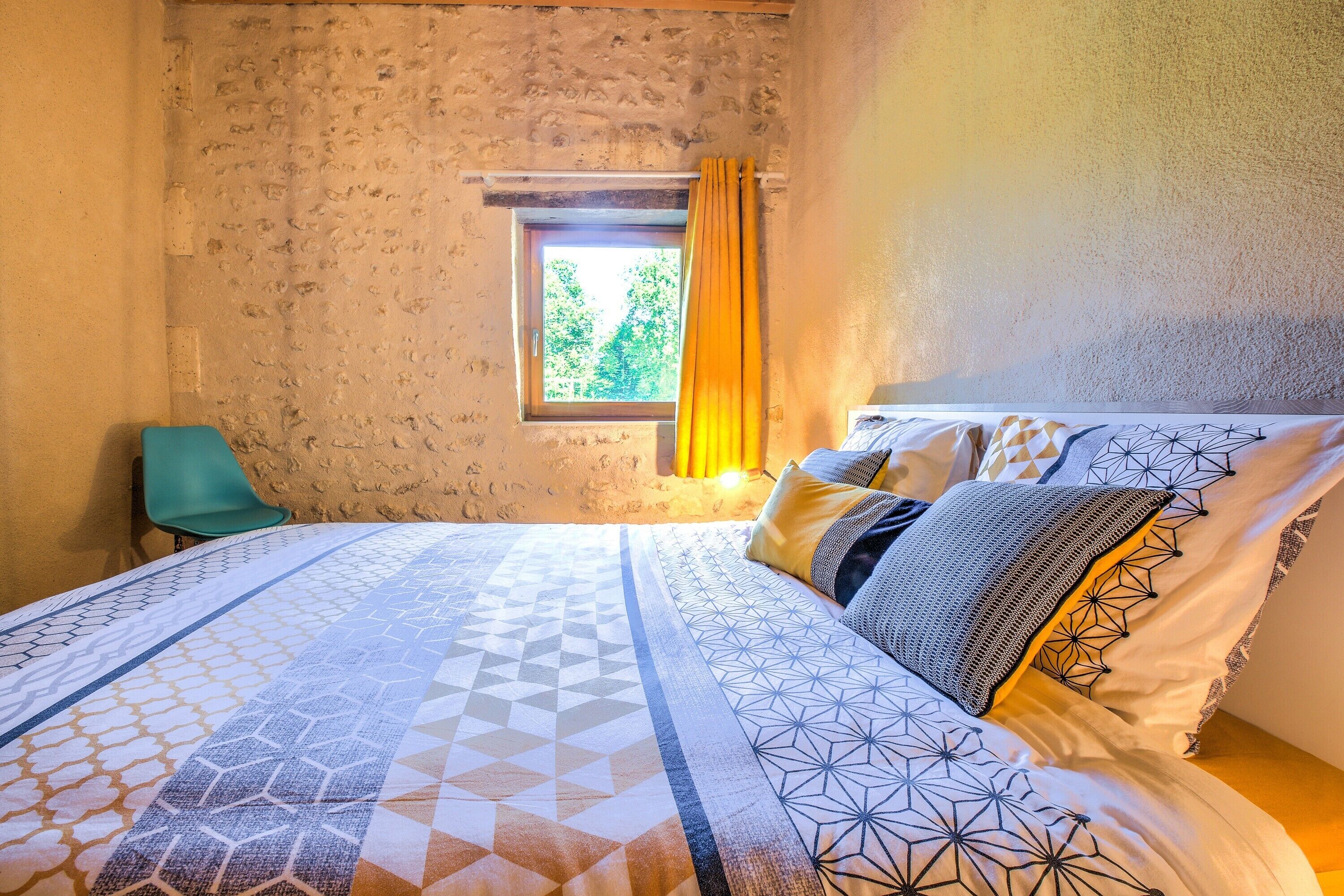A bedroom with a bed, a window with curtains, a chair, and a textured wall.