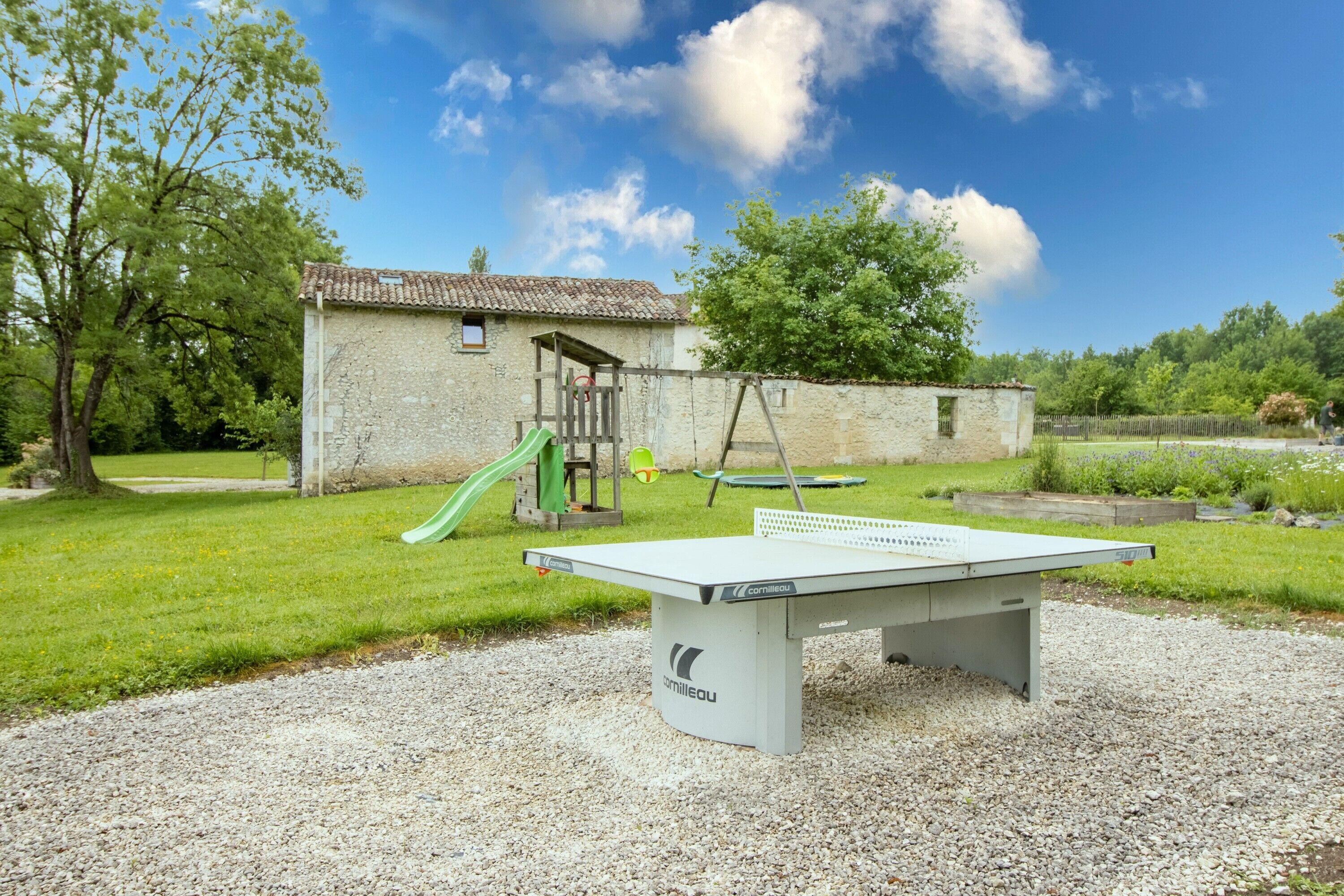 Una mesa de ping pong en un parque, con un edificio de piedra y juegos infantiles al fondo.