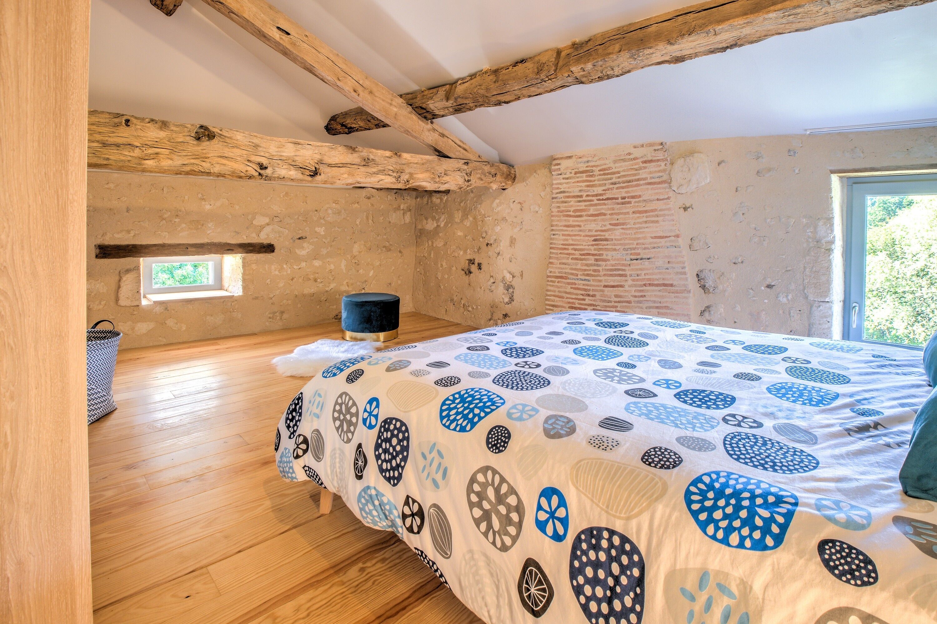 Een slaapkamer met houten vloer, een bed met een geborduurde spreideken, een klein raam en openliggende bakstenen muren.