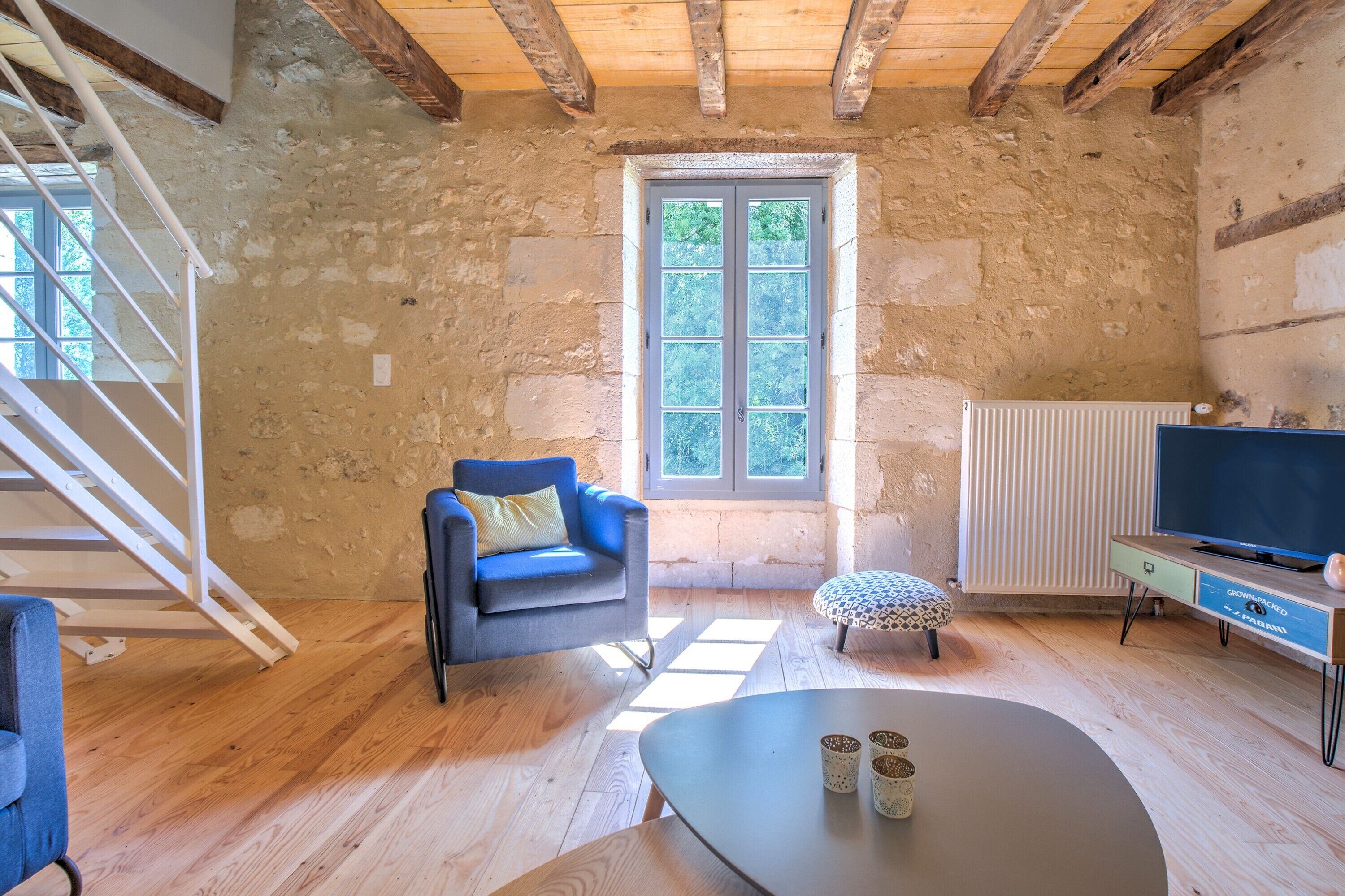 Una stanza rustica con parete in pietra, una scala in legno, una poltrona blu, un tavolo rotondo e un televisore su un mobile in legno.