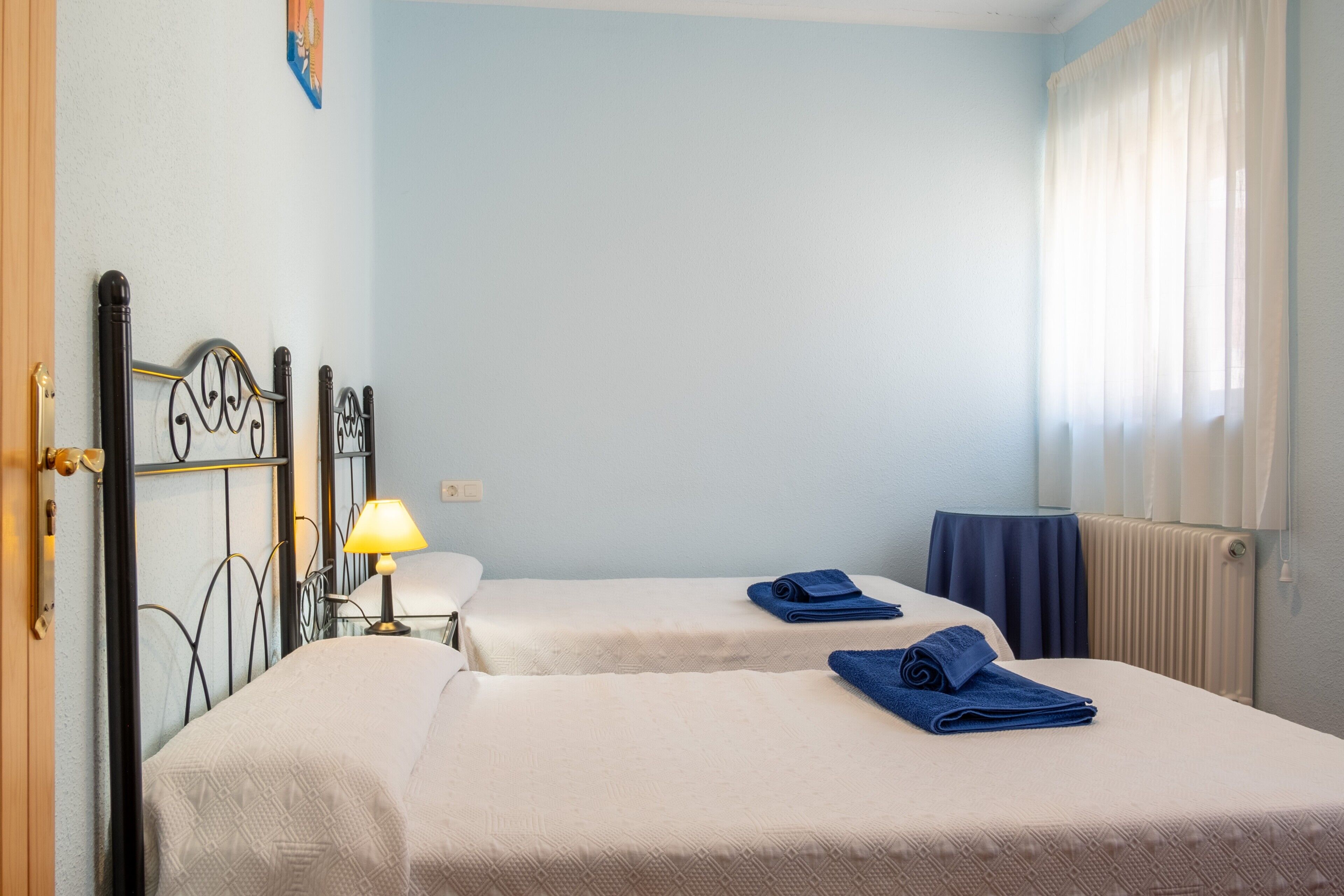 Habitació amb dos llits, una làmpada, un radiador i una finestra amb cortines.