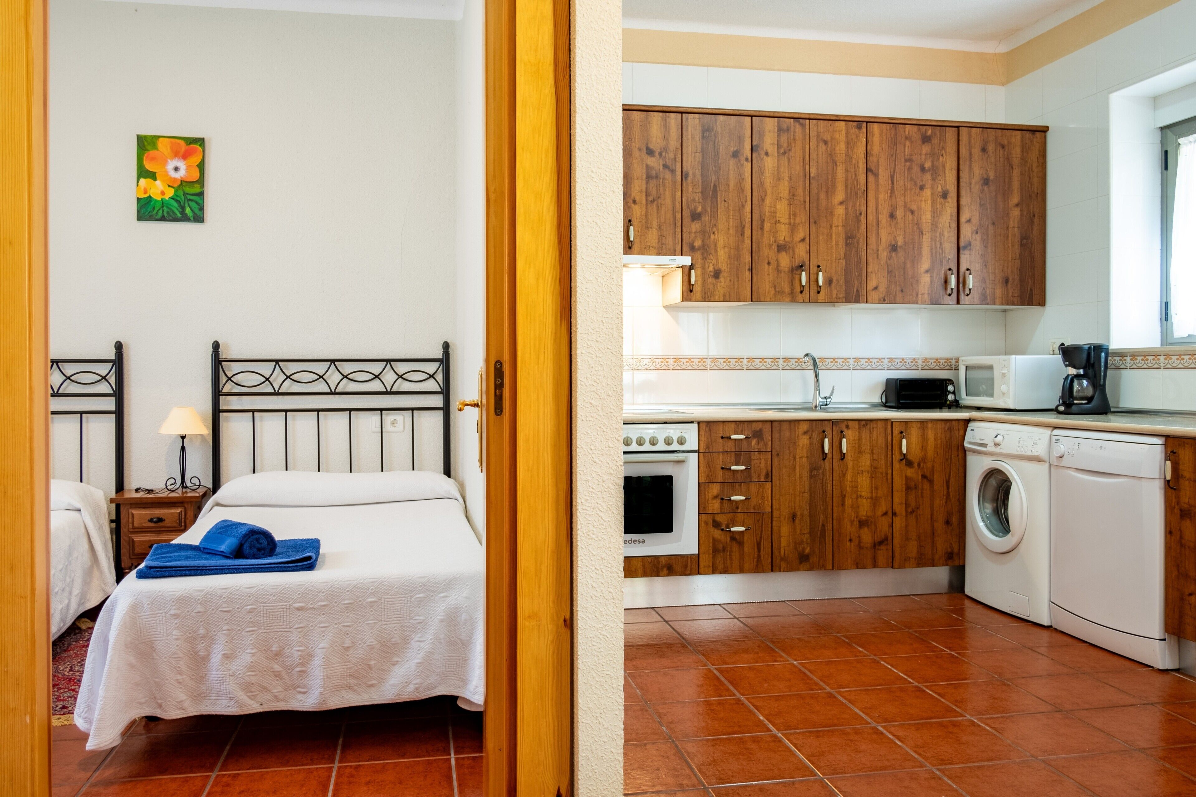A room with a bed, a nightstand, a lamp, and a painting on the wall. Adjacent is a kitchen with wooden cabinets, a microwave, a coffee maker, and a washing machine.