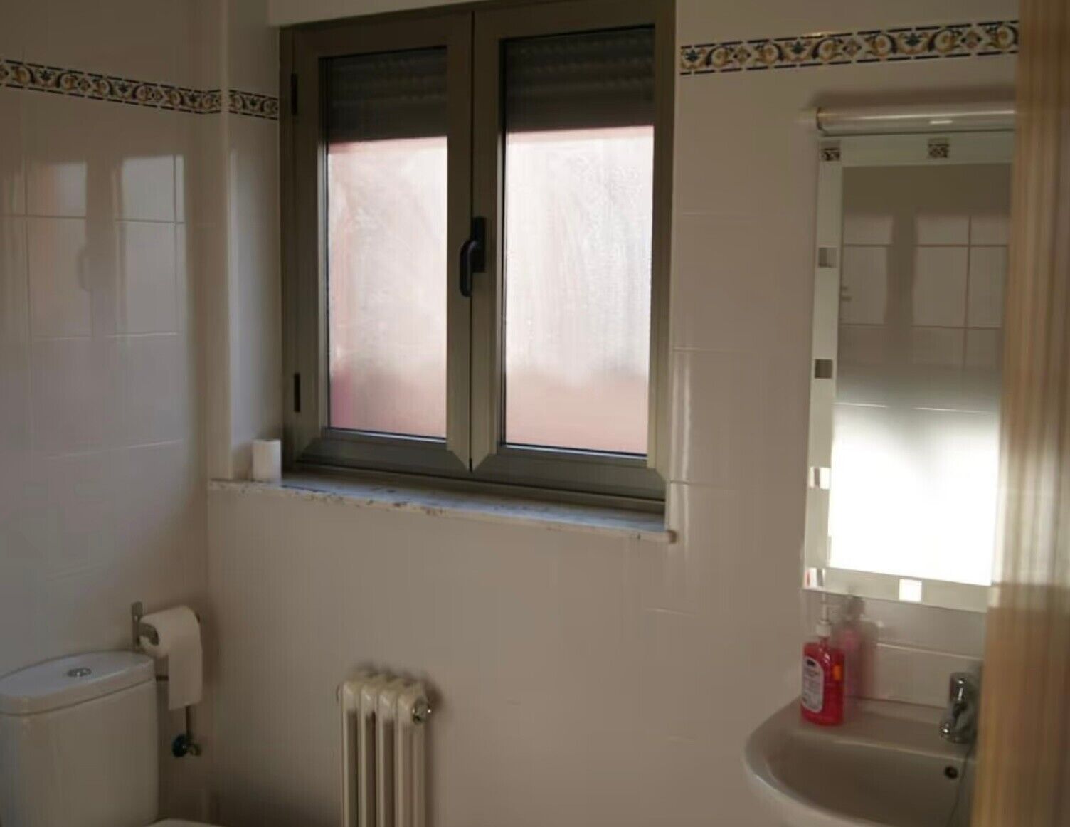 Un baño con inodoro, radiador, lavamanos con espejo y ventana con persianas.