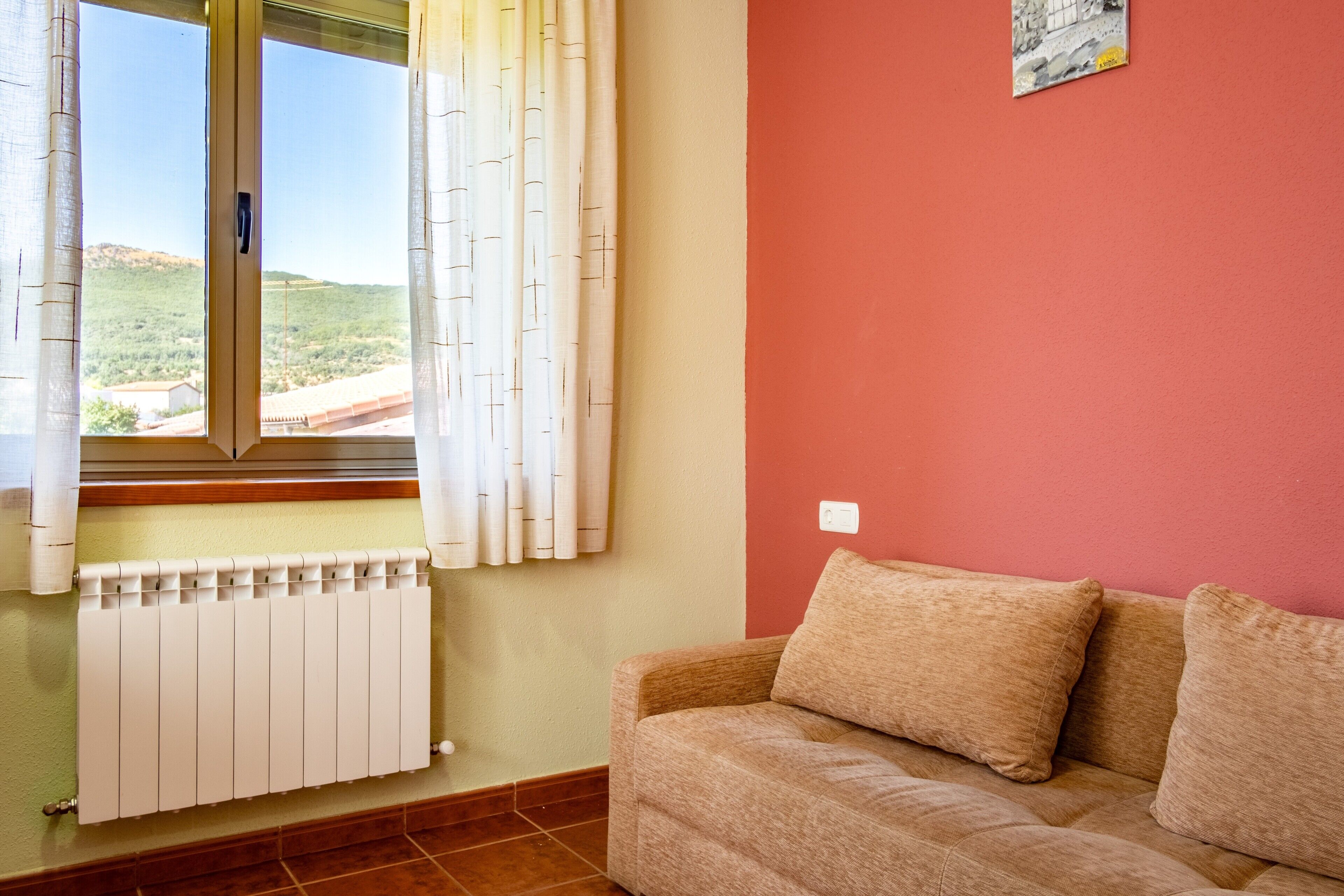 Una habitación con ventana, un radiador, un sofá color crema y una pared roja.