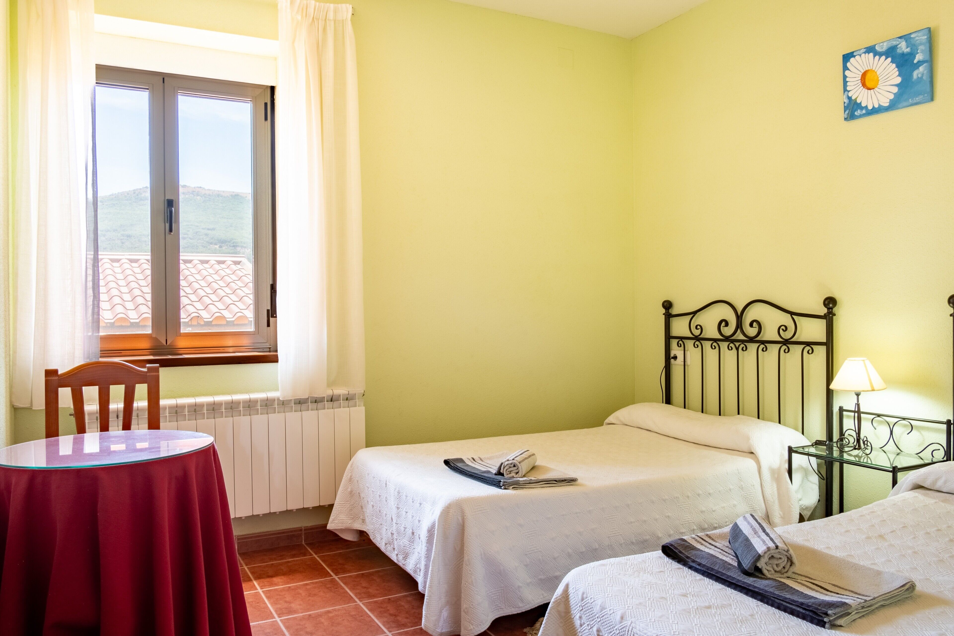 A room with two beds, a table, a window with curtains, a radiator, and a wall painting.