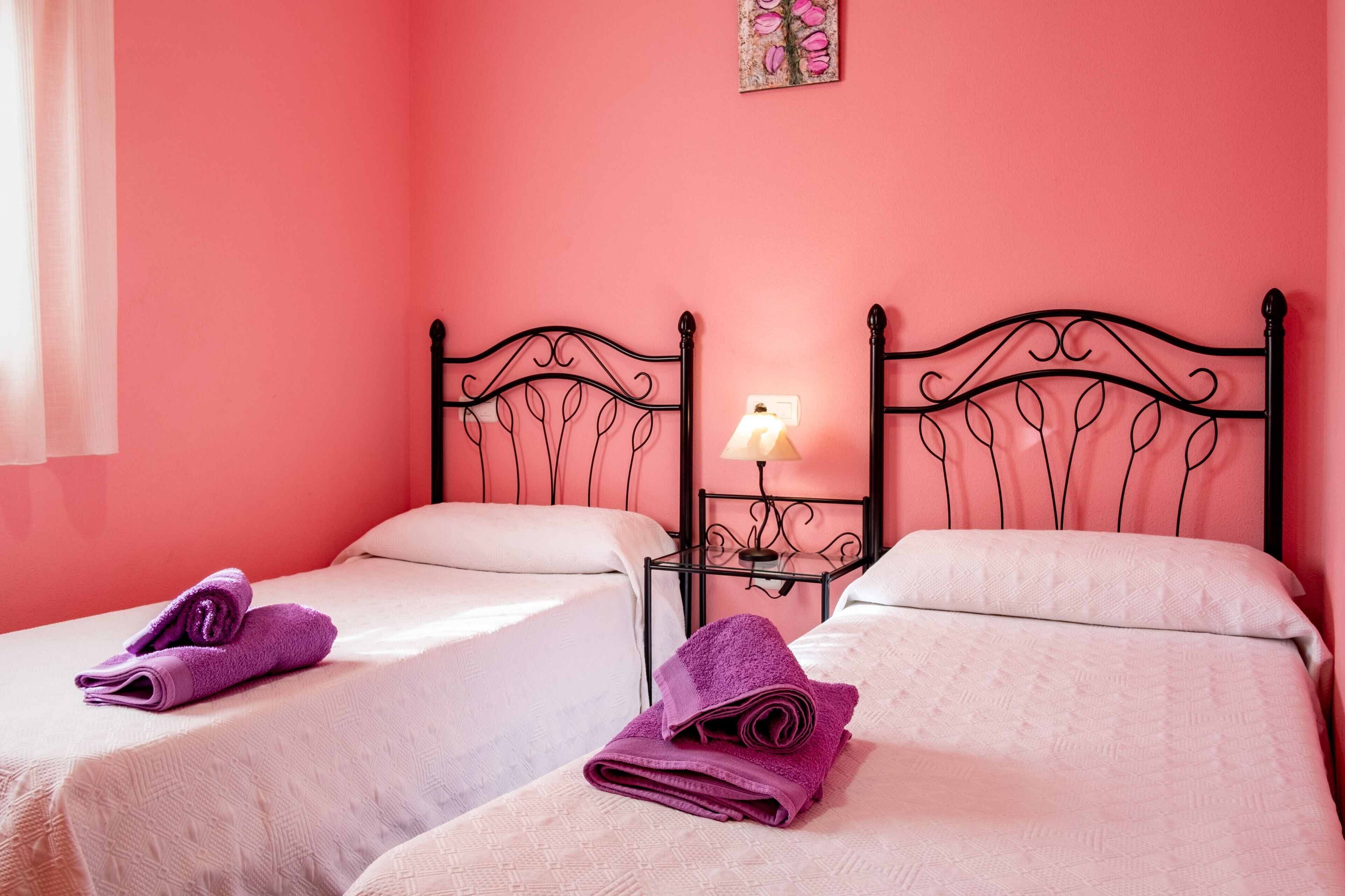 Een kamer met twee bedden, elk met een hoofdbord, een nachtkastje met een lamp, en een opgerolde paarse handdoek op elk bed.