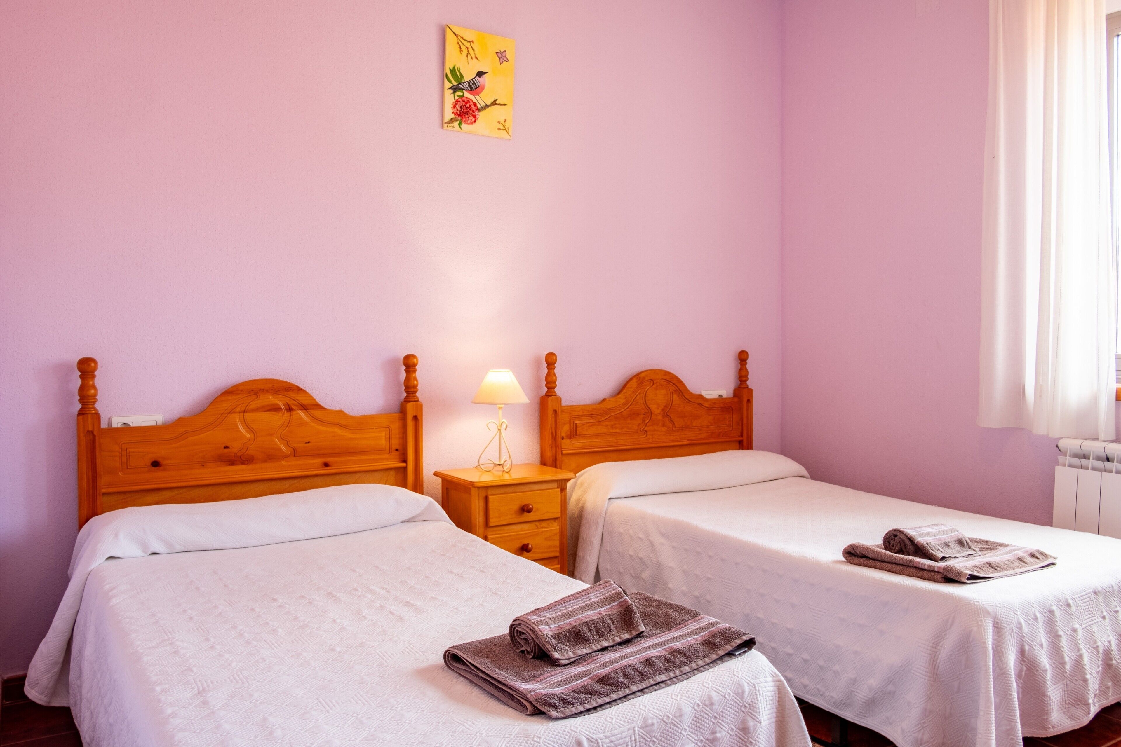 A room with two beds, wooden headboards, a nightstand with a lamp, and a window with curtains.