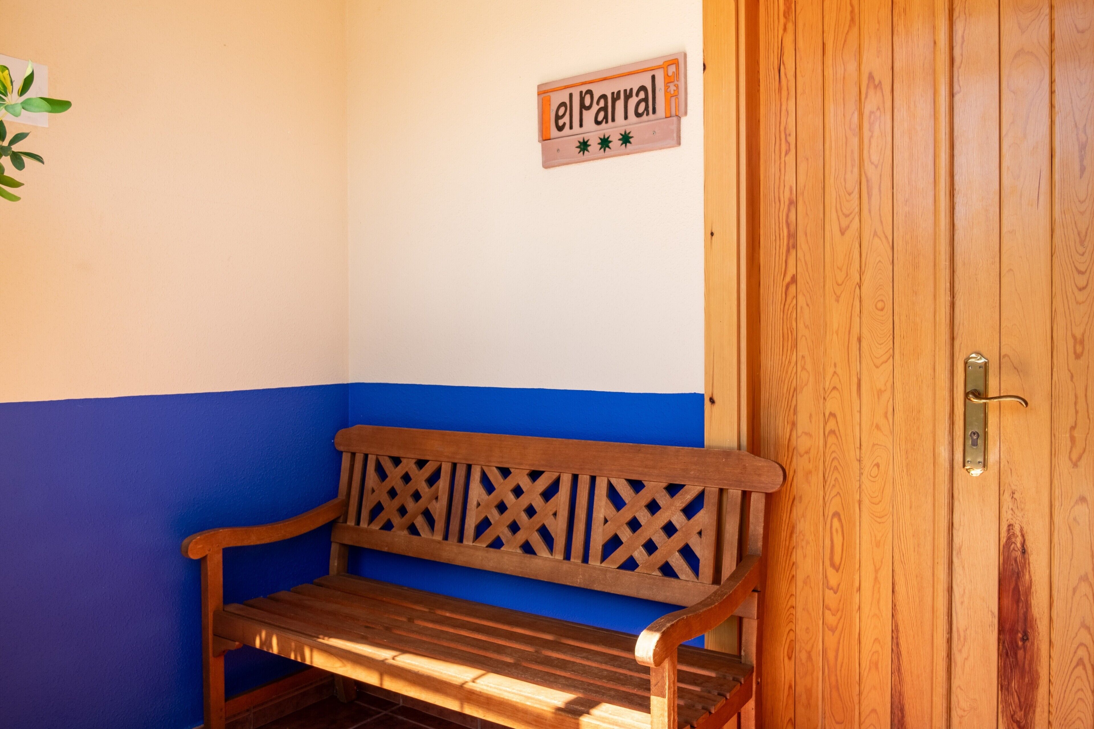 Eine hölzerne Bank vor einer Wand in Blau-Weiß gehalten, eine Tür mit Messinggriffen und ein Schild mit der Aufschrift 'elParral'.