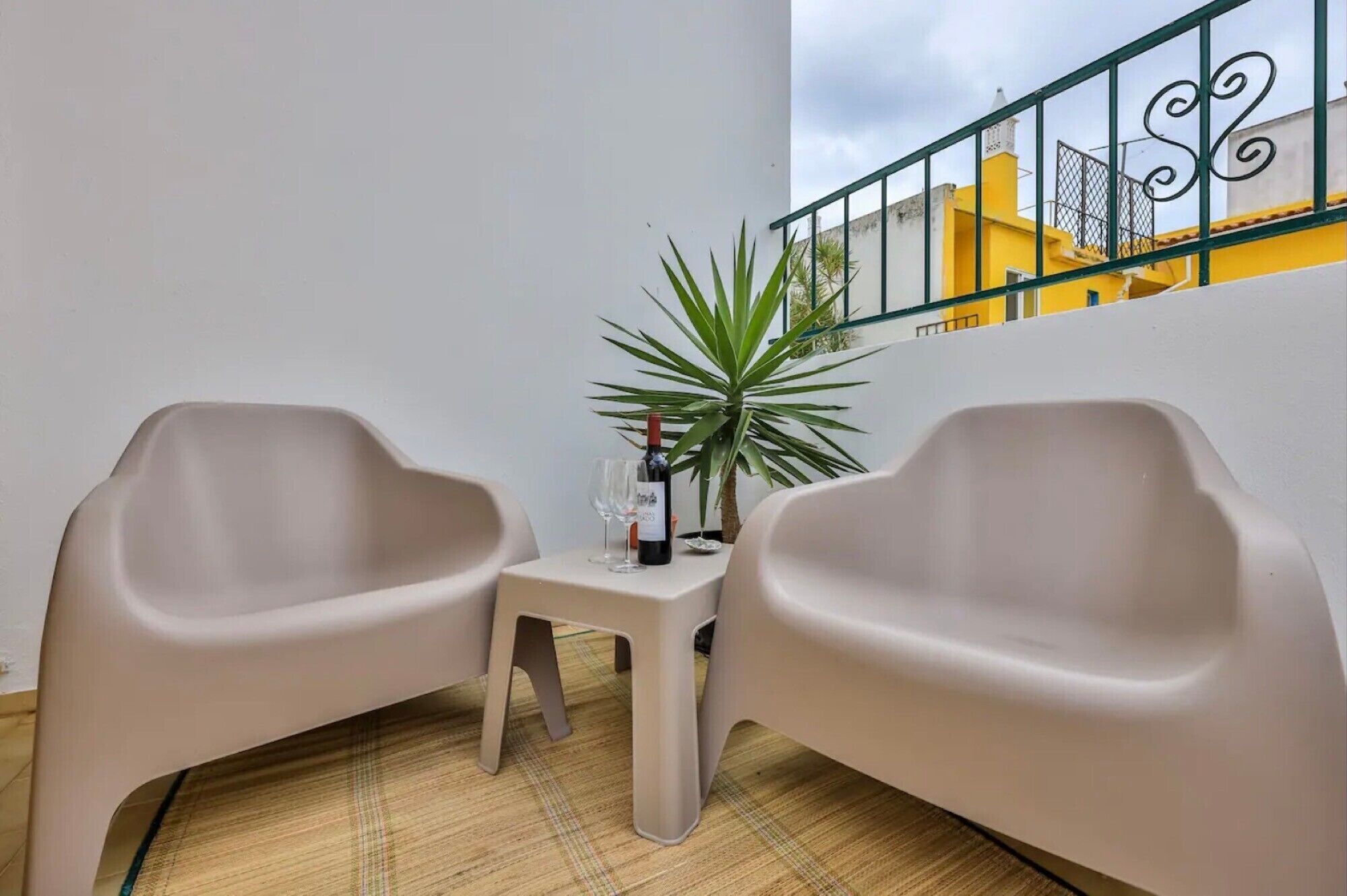 A balcony with two beige chairs, a small table, a potted plant, and a bottle of wine.