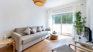 A modern living room with a sofa, a small table with a plant, a wicker lamp, a sliding glass door, and a large potted plant. - Holiday Home 'Casa Cláudia - Cond  Atalaia' with Shared Pool, Private Terrace and Wi-Fi (Lagos)