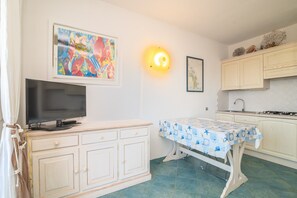 A room with a TV on a cabinet, a table with a patterned tablecloth, a wall-mounted light, and a kitchen area with cabinets.