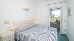 Un dormitorio con cama, mesitas de noche, una lámpara de pared, luz de techo y vistas por una puerta.