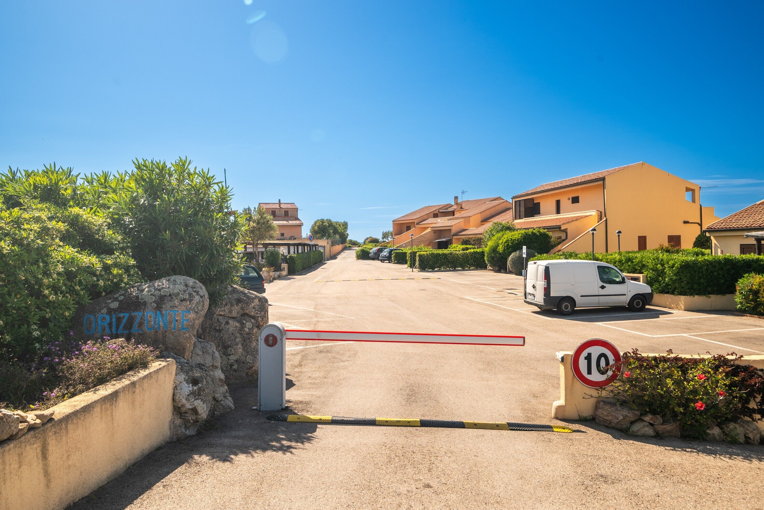 A gated entrance to a residential area with a speed limit sign, a white van parked inside, and a clear blue sky.