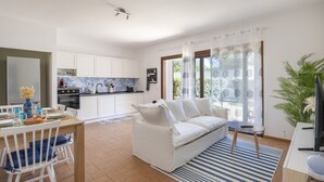 A modern living room with a white sofa, a dining table with chairs, a kitchen in the background, and a large window with curtains. - Apartment 'Flat Arcipelago' with Shared Pool, Wi-Fi and Air Conditioning (Palau)