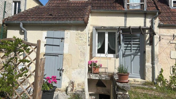 Uma casa de dois andares com telhado em telhas, uma pequena varanda com plantas em vasos e um portão de madeira.