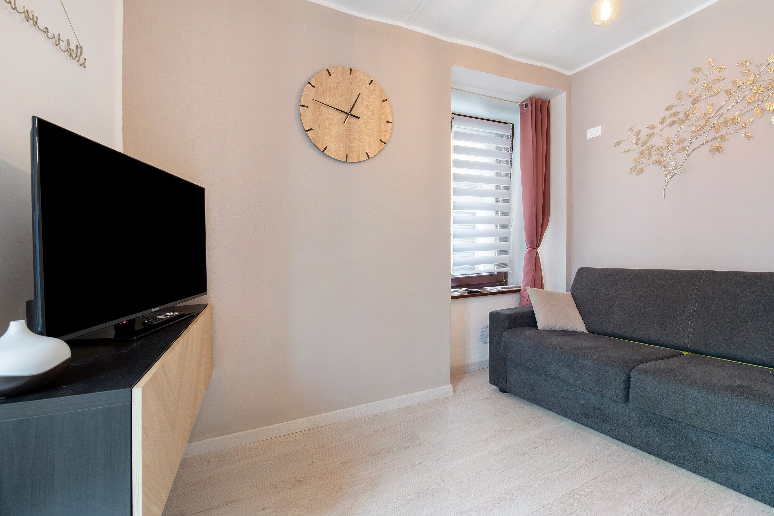 A modern living room with a flat-screen TV, a wooden clock, a grey sofa, and a decorative branch on the wall.