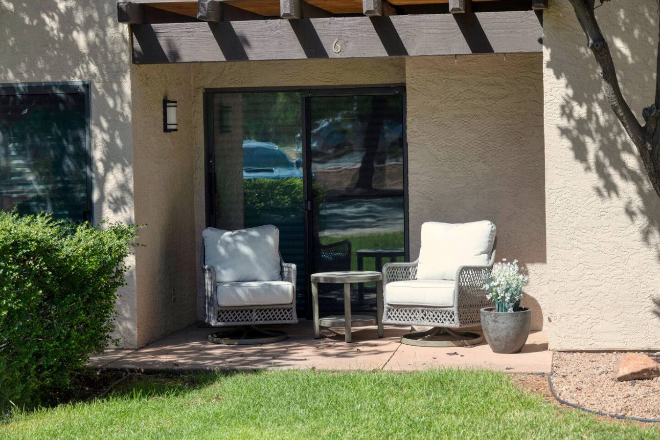 Un patio couvert avec deux fauteuils en osier blancs et une petite table, une plante en pot, et une vue sur une étendue d’eau par une porte vitrée.