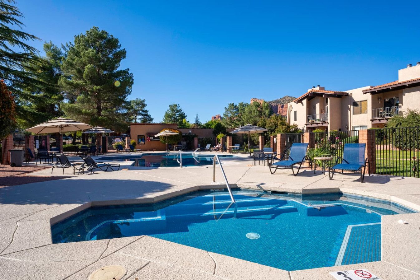 A swimming pool area with lounge chairs and umbrellas, surrounded by buildings and trees.