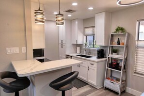 Una cocina moderna con armarios blancos, encimera de mármol, dos taburetes negros, iluminación colgante, una cafetera y un pequeño estante con adornos.