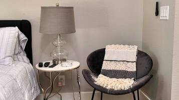 A bedroom with a bed, a bedside table with a lamp, a chair with a throw blanket, and a wall socket.