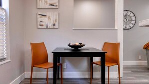A dining area with a black table and two orange chairs, a bowl of fruit on the table, and abstract art on the wall.