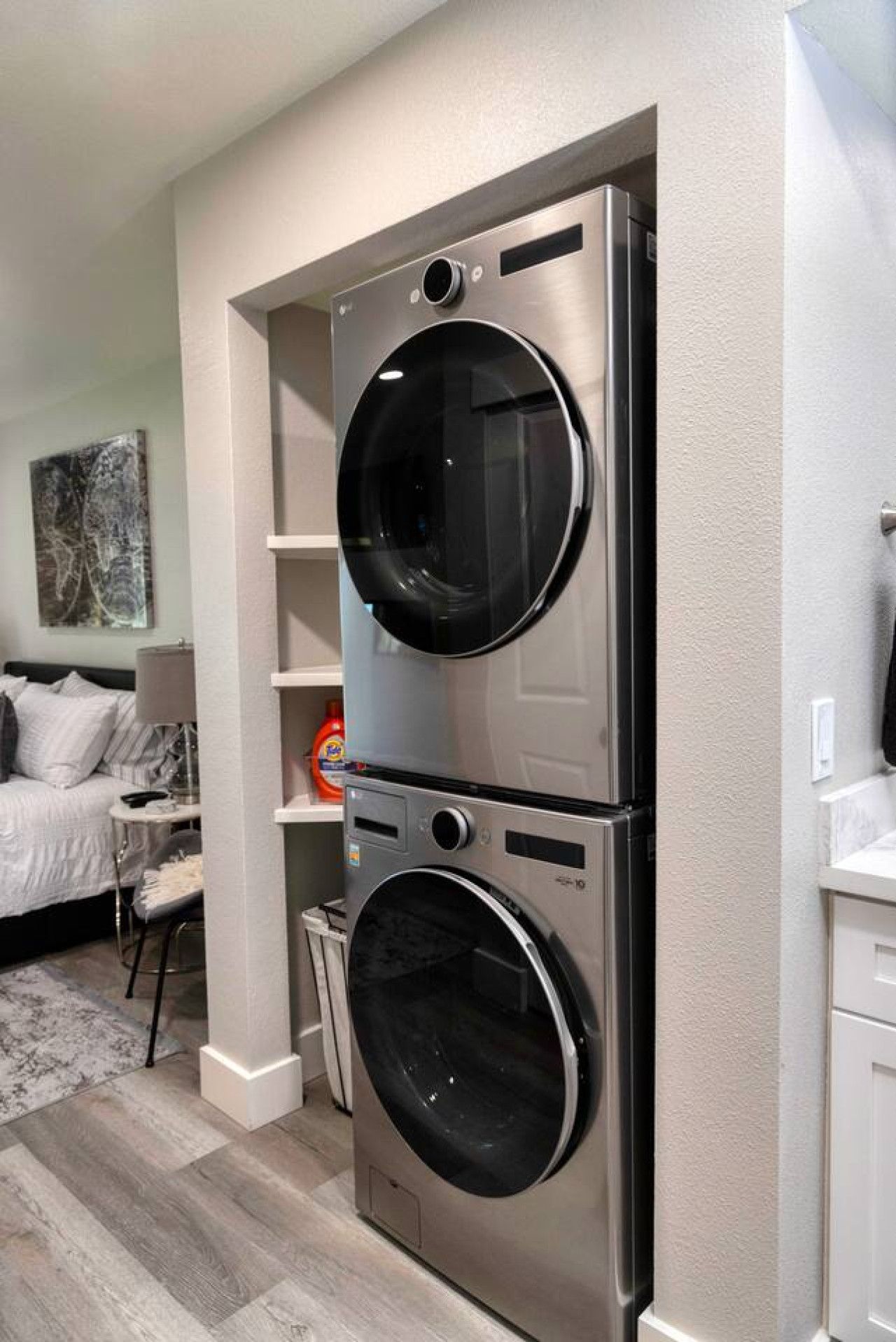 A compact laundry room with a double stainless steel washer and dryer, a small shelf unit, and a bed with a nightstand in the adjacent bedroom.
