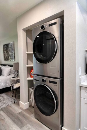 A compact laundry room with a double stainless steel washer and dryer, a small shelf unit, and a bed with a nightstand in the adjacent bedroom.