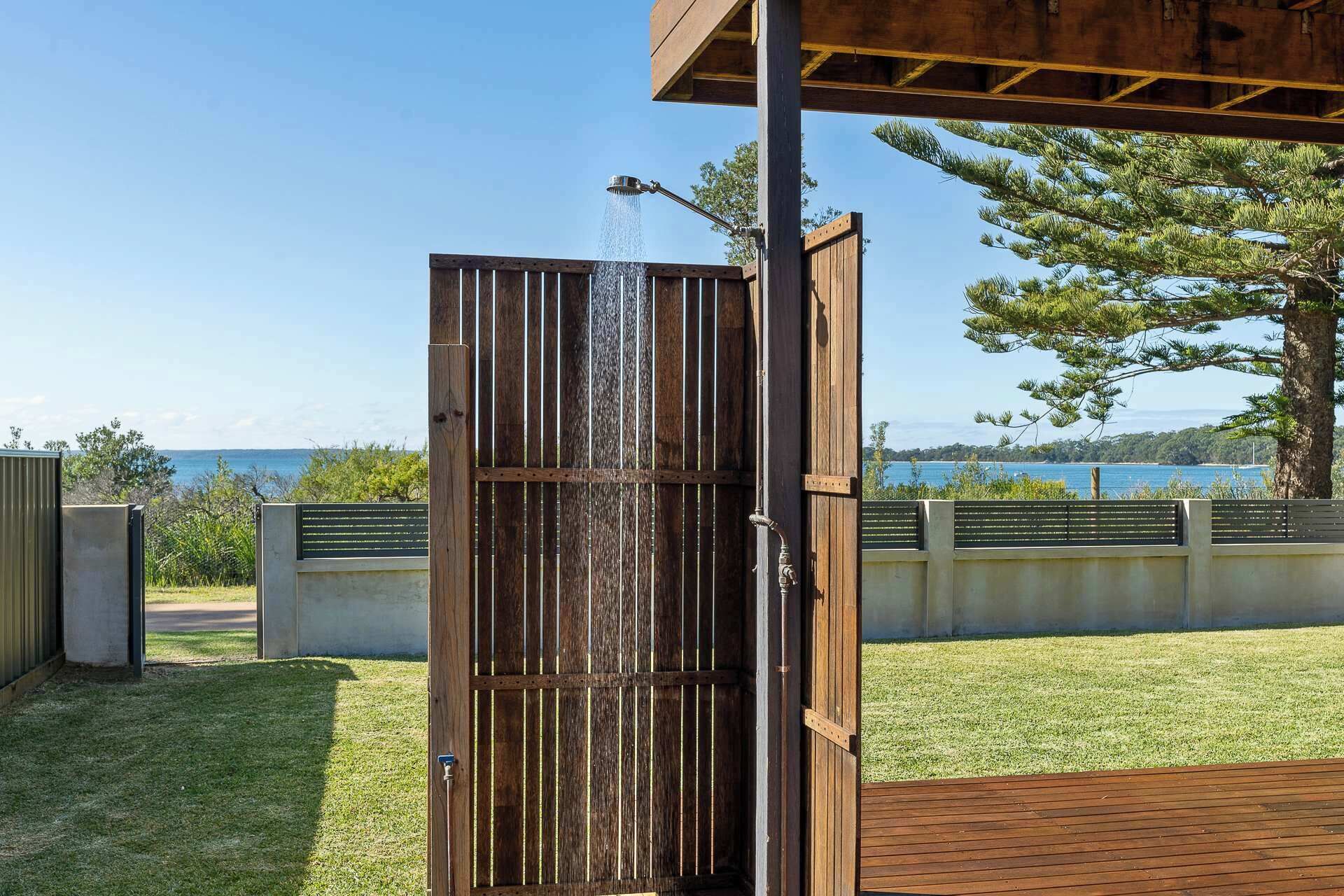 Ducha exterior de madera con agua corriendo, frente a un cielo despejado y una masa de agua.