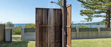 Ducha exterior de madera con agua corriendo, frente a un cielo despejado y una masa de agua.