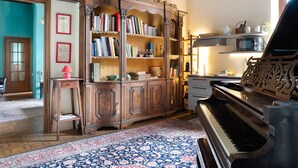 A room with a grand piano, a wooden bookcase, a patterned rug, and a kitchen area with modern appliances. - Apartment 'Il Flauto Magico' with Mountain View, Shared Garden and Wi-Fi (Serralunga d'Alba)