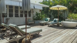A wooden deck with lounge chairs and umbrellas, surrounded by tropical plants and a building with a tiled roof. - VILLA BELLA SESEH (Cemagi)