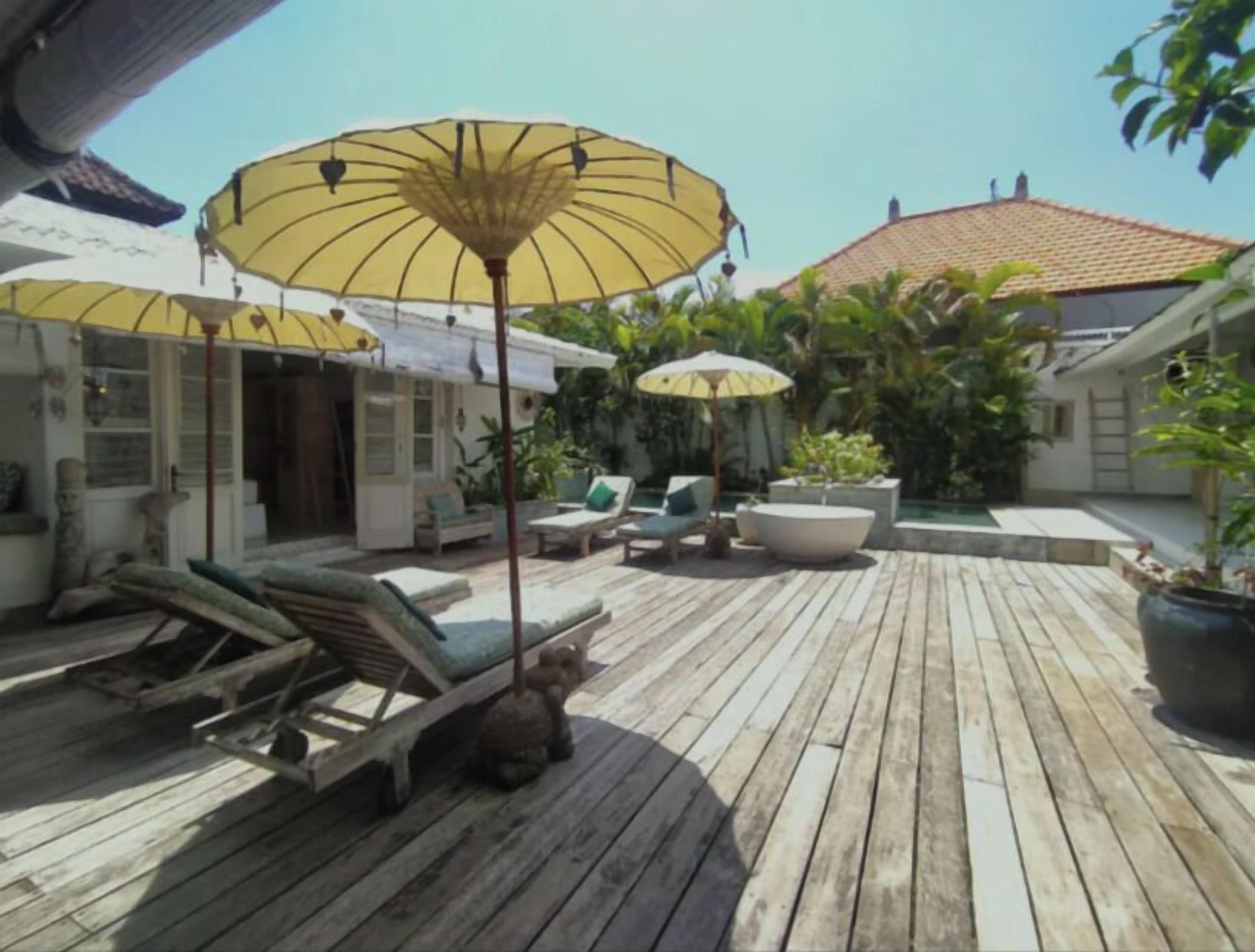 A wooden deck with lounge chairs and umbrellas, surrounded by potted plants and a swimming pool.