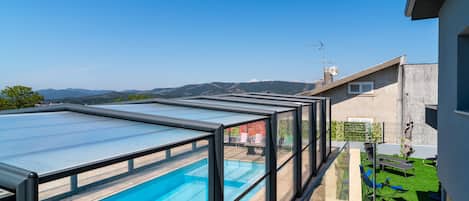 A rooftop swimming pool with glass walls, a patio area with outdoor furniture, and a view of distant mountains.