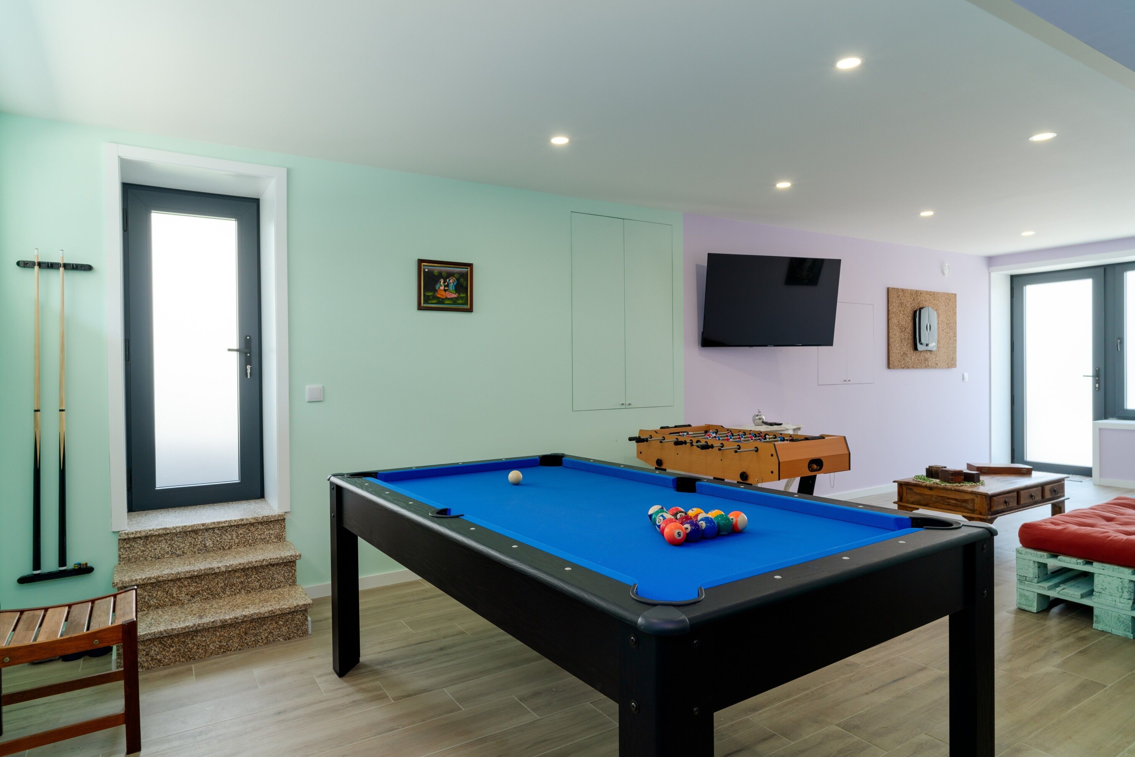 A pool table with balls, a foosball table, a television mounted on the wall, and a staircase leading to an upper level.