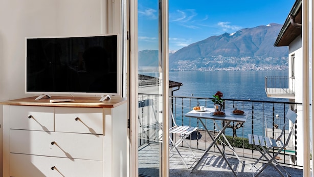 A balcony with a TV, a table with a flower vase, and a scenic view of a lake and mountains. - Garden on the Lake 1 in Vira Gambarogno, Gambarogno, Vira, Switzerland (Gambarogno, Vira)